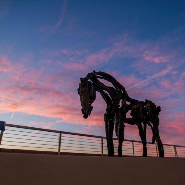 A horse sculpture is seen along a railing against a pink sky.